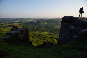 Curbar edge evening This landscape photograph captures Curbar Edge in the evening during spring. The image showcases the rugged rock formations of Curbar Edge, a well-known landmark in Derbyshire, United Kingdom, overlooking the lush, green rural scenery of the Peak District. Nature is prominent in the scene, with rolling fields, wooded areas, and distant hills bathed in soft, low sunlight. A person stands on top of Curbar Edge, emphasising the scale of the natural landscape. The photograph illustrates the tranquil beauty of the rural Peak District in Derbyshire, highlighting one of the United Kingdom's iconic nature destinations.